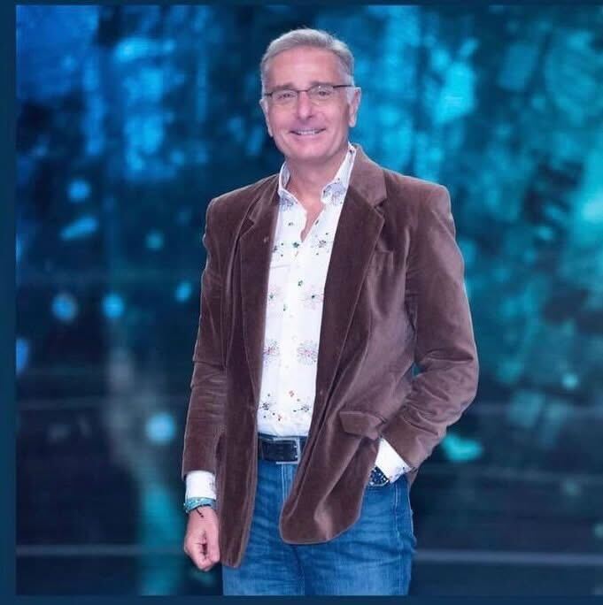 ブラウンのコーデュロイジャケットに花柄シャツとデニムを合わせた男性。ブルー系の背景の中で笑顔を見せる、大人のカジュアルスタイル。