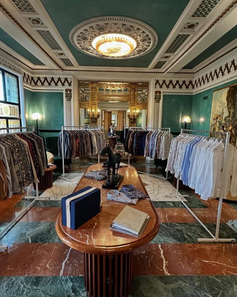 イタリアの重厚なクラシック建築のショールームに並ぶ華やかな柄シャツのコレクション。天井のシャンデリアと大理石の床が高級感を演出している