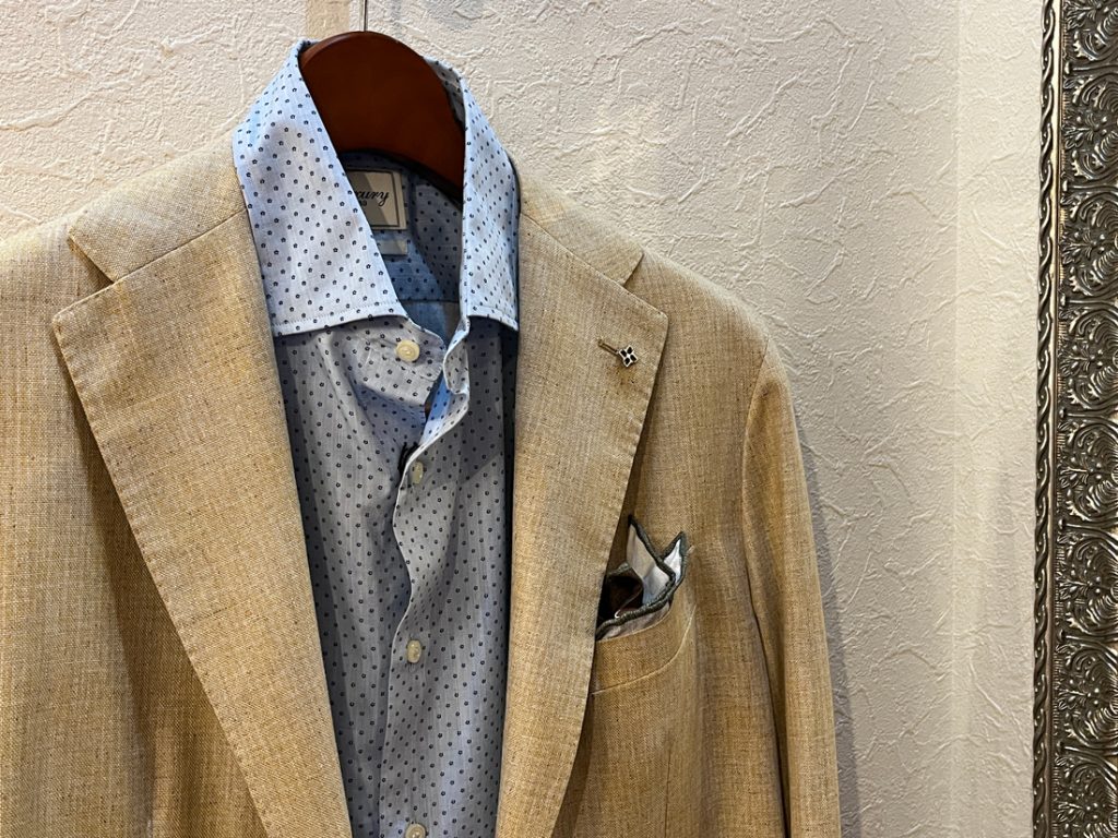 ベージュのジャケットに水色のドット柄シャツを合わせた上品なコーディネート。胸元にはポケットチーフとラペルピンを添え、エレガントな印象に仕上げている。