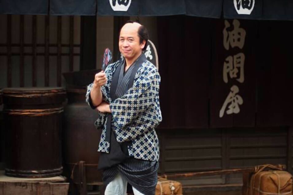 江戸時代の町人風の装いをした男性。幾何学模様の着物に身を包み、扇を手に笑みを浮かべている。背景には商家風の建物と暖簾が見える。