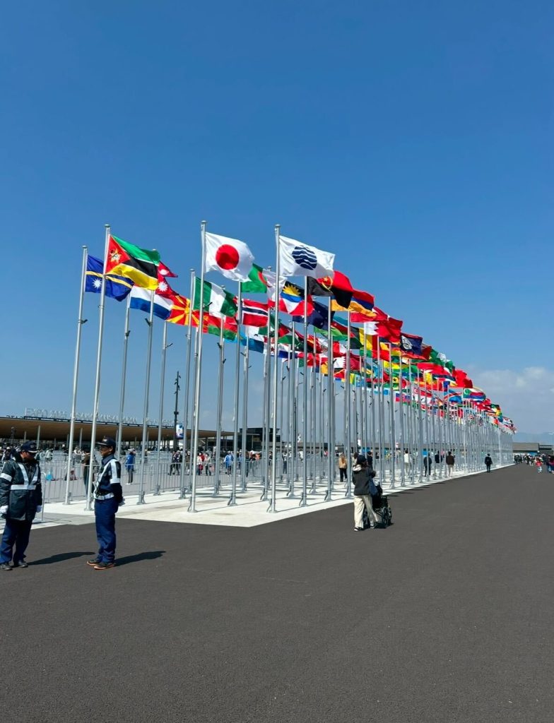 大阪・関西万博の会場入り口に掲げられた各国の国旗と、青空のもとを歩く来場者たちの様子