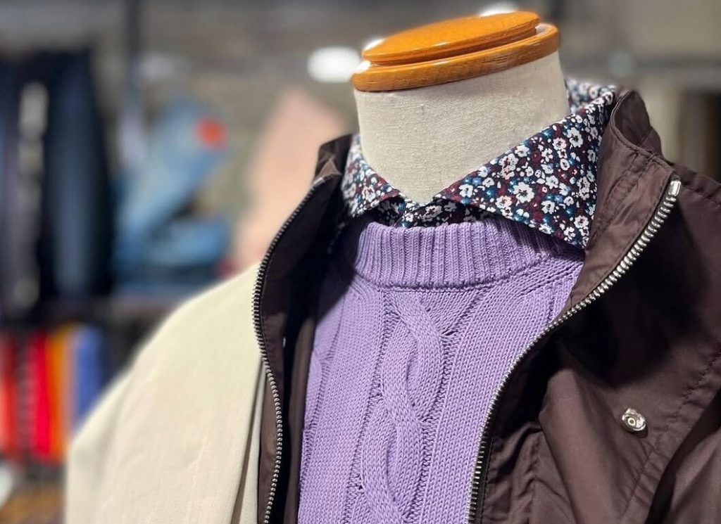ラベンダーのケーブルニットと、花柄シャツ、ブラウンのブルゾンを重ねたメンズ春コーデ。肩にはベージュのパンツをコーディネート。