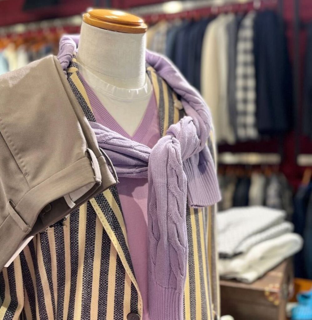 春の気温差に対応するイタリアメンズスタイル。ラベンダーカラーのニットを肩掛けし、ストライプジャケットとVネックニットで上品に仕上げたレイヤードコーデ。