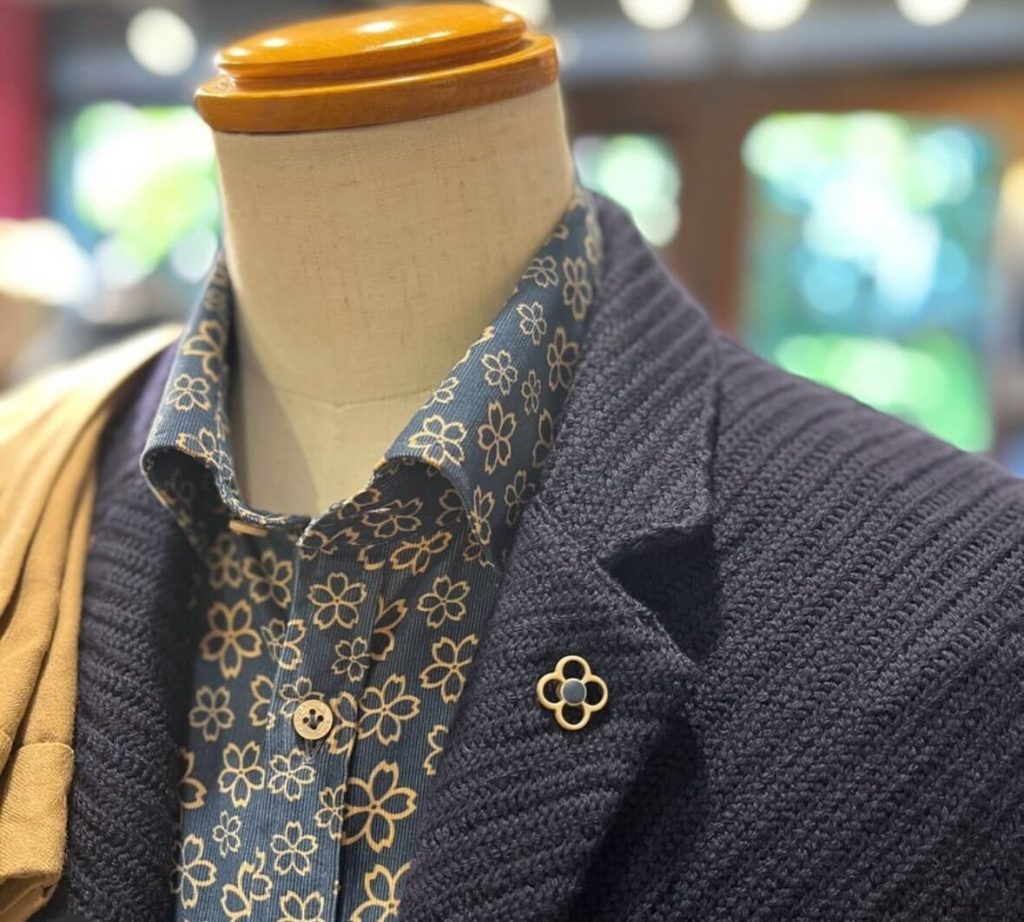 ネイビーのニットジャケットに、花柄のシャツを合わせたメンズコーディネート。胸元にはゴールドのフラワーモチーフピンがアクセントに。