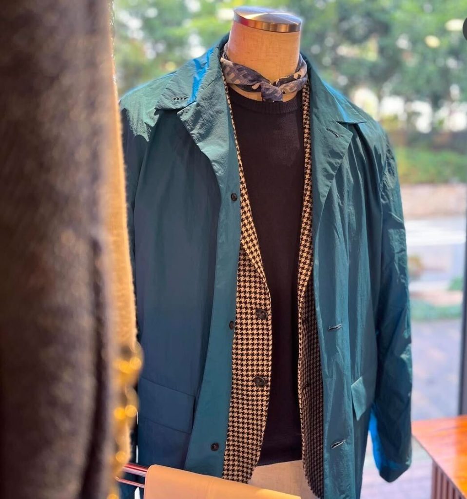 千鳥格子のジャケットとブラックニットの上に、ブルーグリーンの軽量ステンカラーコートを重ねたイタリア春スタイル。3月の防寒と春の先取りを両立する大人のコーデ。