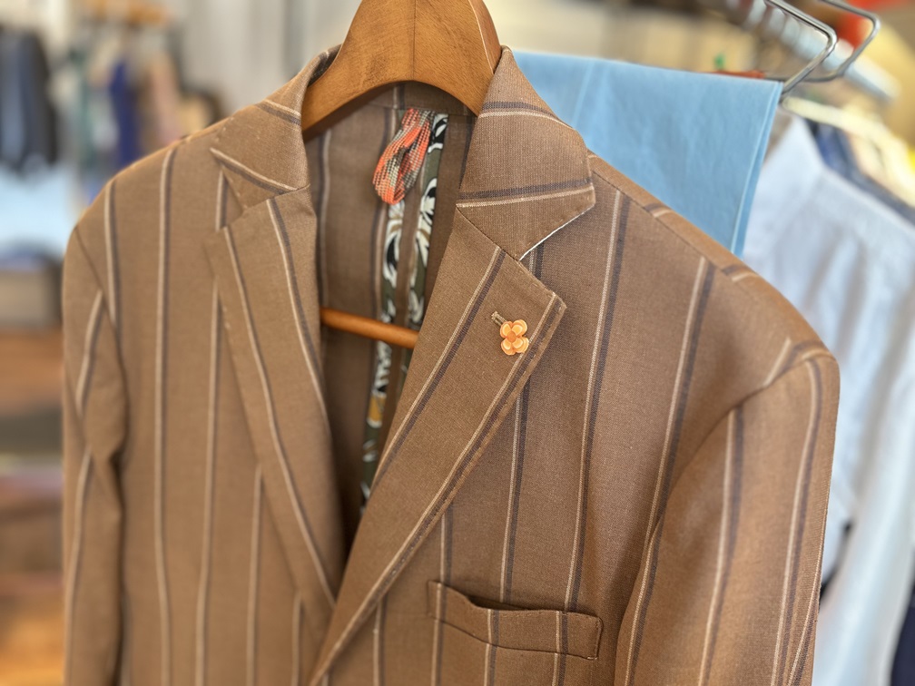 SSEINSEのストライプジャケットのラペルに飾られたオレンジ色の花型ピン。遊び心あるディテールが印象的。