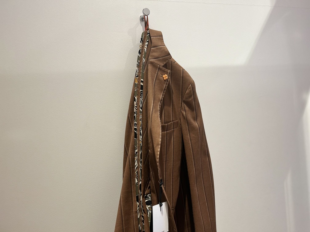 SSEINSEのリネン混ストライプジャケット。壁に掛けられた状態で、オレンジのラペルピンと内側の柄パイピングが見えるディテールカット。春夏の軽やかなメンズジャケットスタイルに最適。