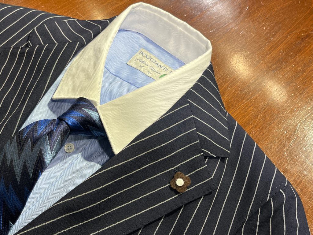 ネイビーのストライプスーツに、白いクレリックカラーのシャツとブルー系のジグザグ柄ネクタイを合わせたクラシカルな装い。襟元には花型のラペルピンが添えられており、洒落感を引き立てている。