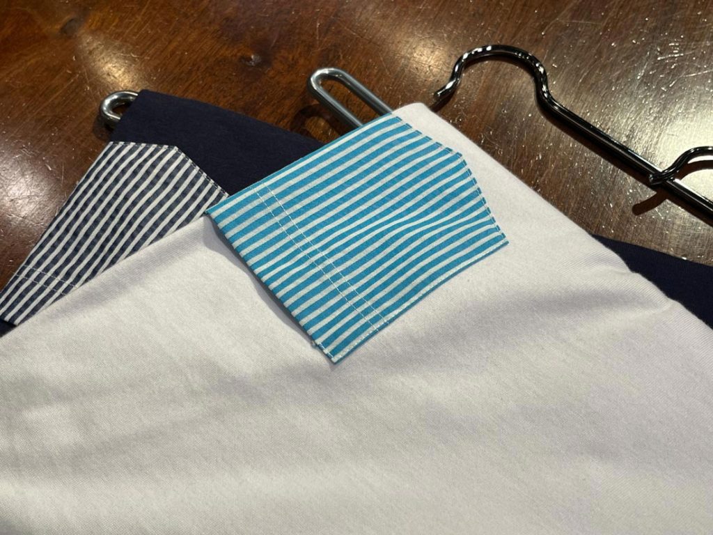 白地のTシャツにブルー系ストライプ柄のポケットがデザインされたディテール。袖にもストライプ切り替えが施され、個性的な配色が目を引く。