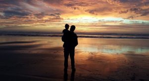 夕暮れの海辺で子どもを抱く父のシルエット。親子の絆と人生の有限性を象徴する場面。