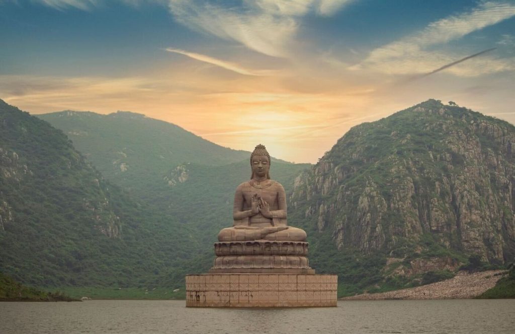 湖の中央にたたずむ巨大な仏像。背後には緑豊かな山々が連なり、夕焼けの空が神聖な雰囲気を演出している。