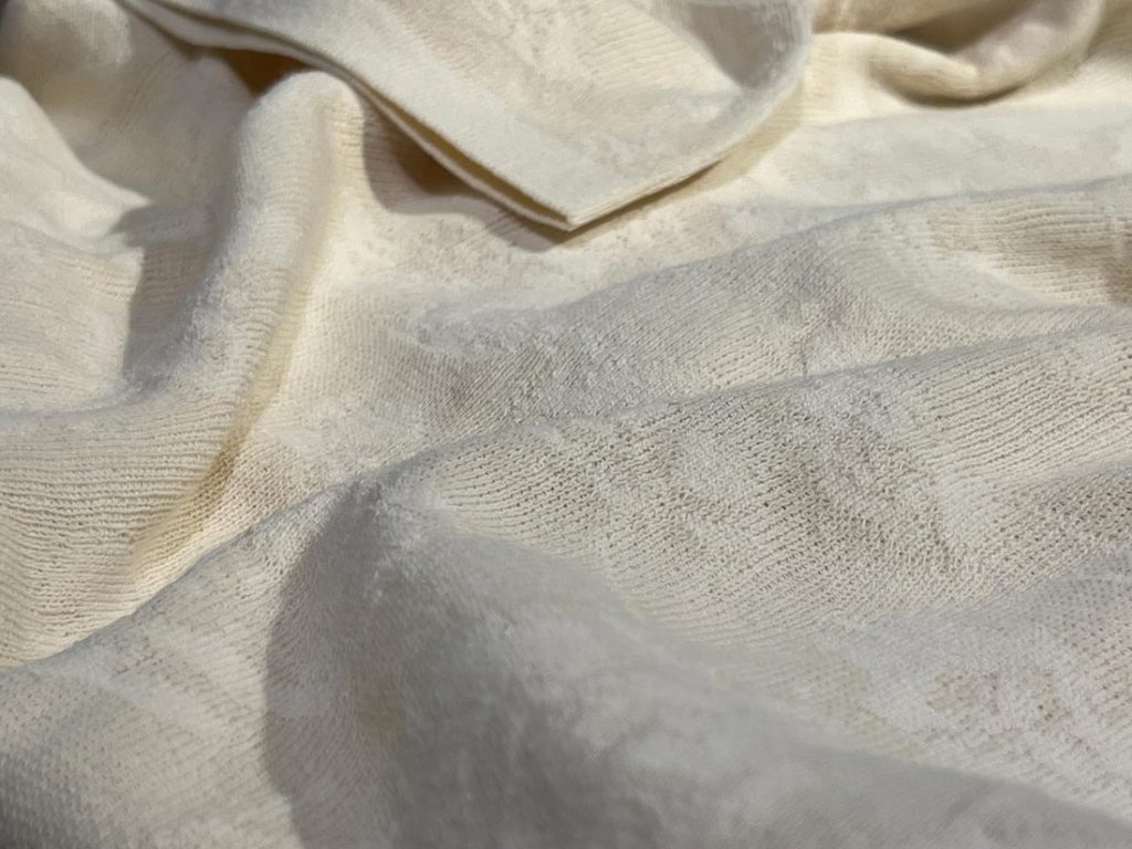 クリーム色のジャカード柄ポロシャツ生地をクローズアップ。繊細な柄としなやかなストレッチ感が手触りまで伝わる質感表現。