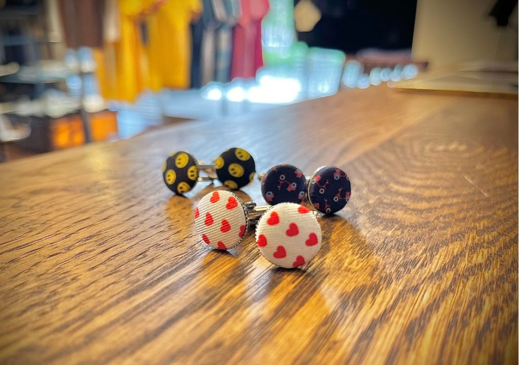 FEFE（フェフェ）のカフスボタン3種。赤いハート柄、スマイリーフェイス柄、赤いスクーター柄がウッドテーブルの上に並び、ユニークで遊び心あるデザインが魅力。