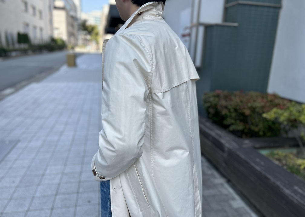 2月の冷たい風を受けて襟を立てたベージュのスプリングコート後ろ姿、春の光が差す街中スタイル